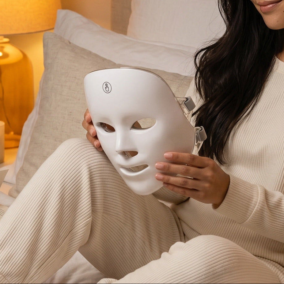 Woman sitting on a bed holding a white face mask in a cozy bedroom setting.
